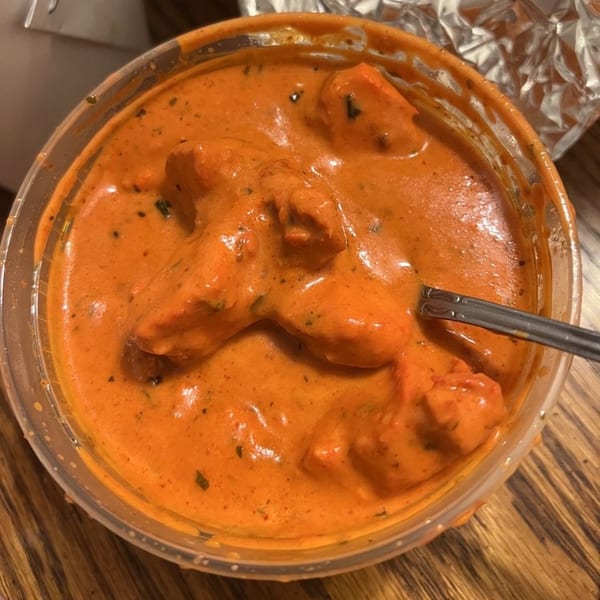 Close-up of Indian butter chicken in a plastic takeout container with a spoon.