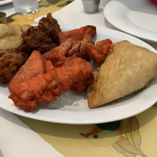 A white plate holds various Indian appetizers: brown pakoras, red chicken pakoras, and a large samosa.